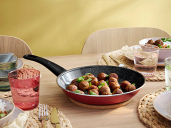 Sartén roja con albóndigas vegetales y hierbas frescas sobre un salvamanteles de corcho en una mesa de comedor de madera preparada para comer.