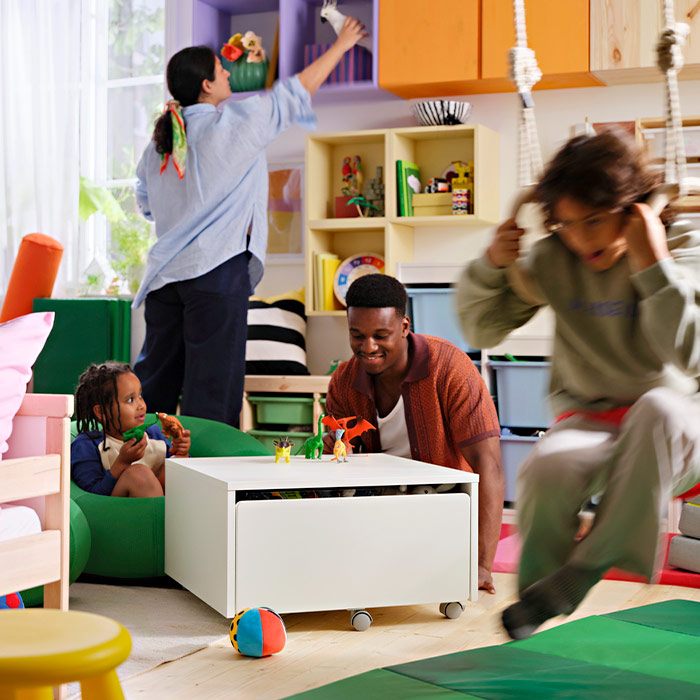 A family enjoys a colorful living room. A man plays with dinosaurs on a white table with wheels while children play around in a bright atmosphere.