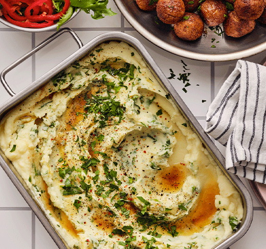 Vista superior de una fuente metálica con puré de patatas cremoso, decorado con mantequilla derretida, pimienta negra y hierbas verdes frescas.