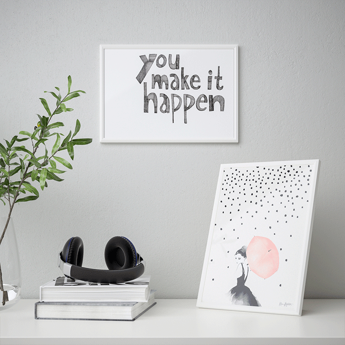 Espacio de trabajo minimalista con un cuadro blanco con la frase "You make it happen" en la pared y una lámina apoyada de una chica con paraguas rosa, junto a auriculares y libros.
