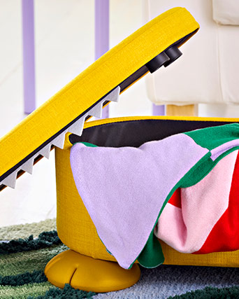 Yellow storage chest with a playful tooth-lined lid and round feet, holding colorful textiles in a neat and tidy corner of the home.
