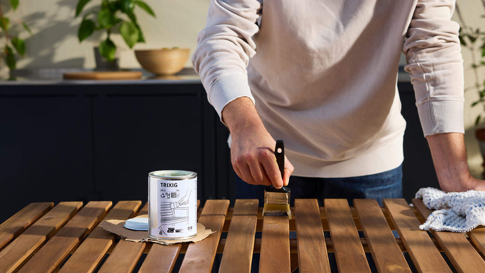 A person is re-staining a wooden outdoor table with a brush next to a can of TRIXIG outdoor wood stain.