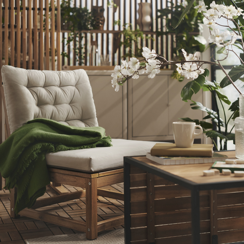 A balcony with greenery features a light brown stained NÄMMARÖ easy chair with light grey-beige cushions and a wooden table.