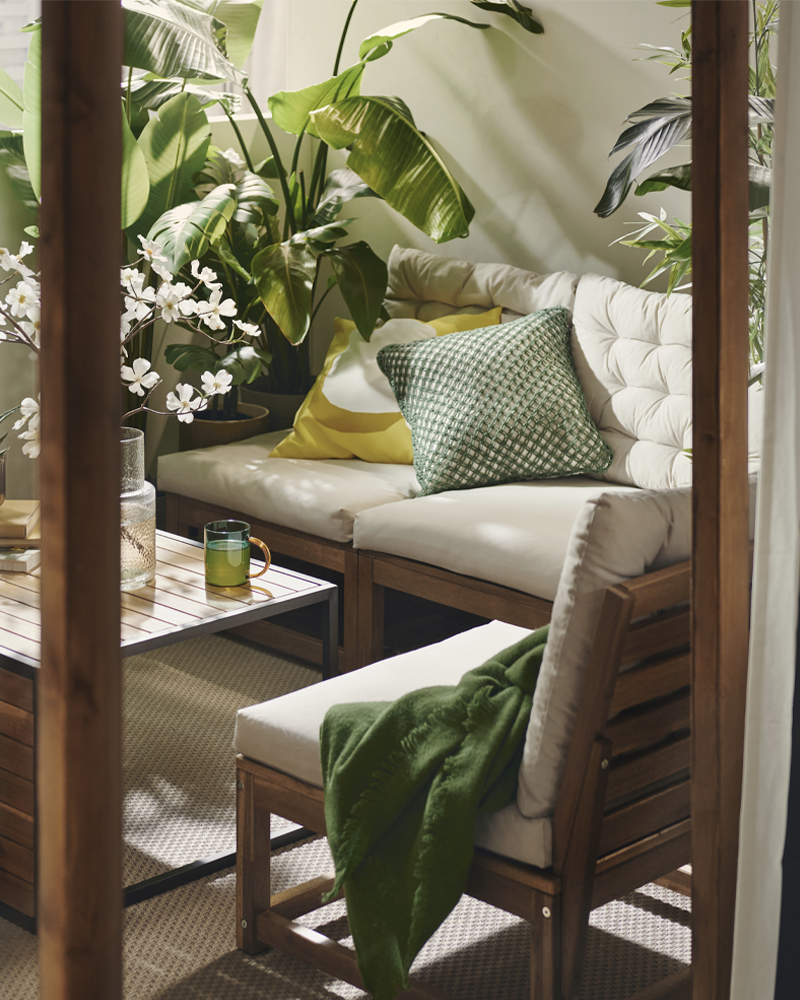 A lush balcony lounge setting with a solid wood NÄMMARÖ modular sofa and a coffee table, colourful pillows and a throw.