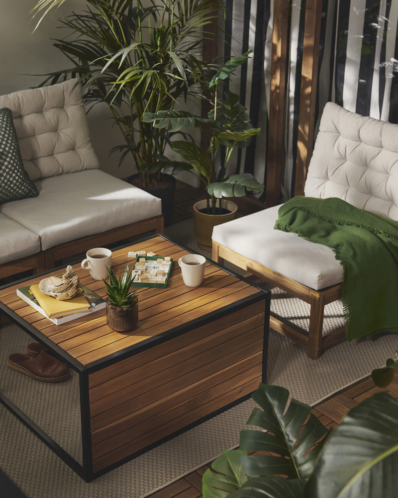 A lush balcony with a versatile light brown HÅKANSKÄR coffee table and seating surrounded by potted plants and soft cushions.