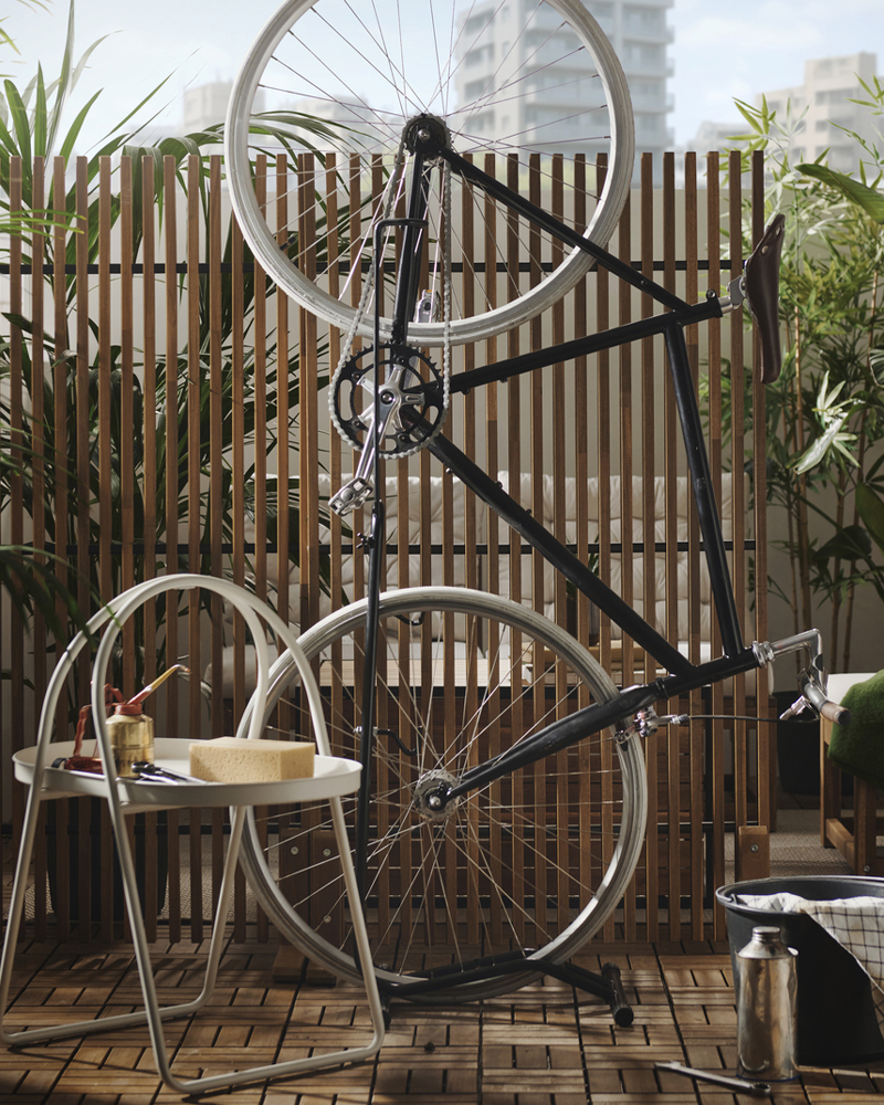 A lush city balcony features a light brown stained NÄMMARÖ privacy screen, surrounded by outdoor furniture and a bike.