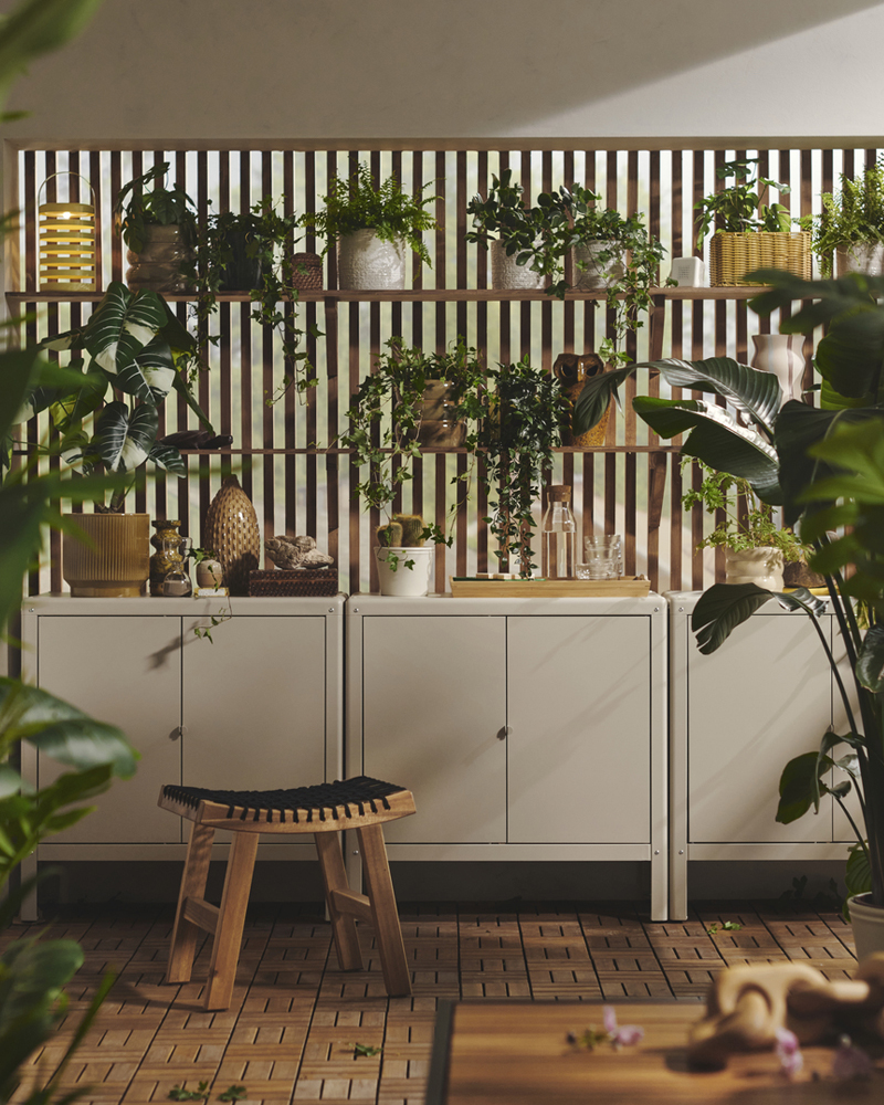 A bright balcony with three beige KOLBJÖRN cabinets, wooden wall shelves displaying various plants on a wall behind it.