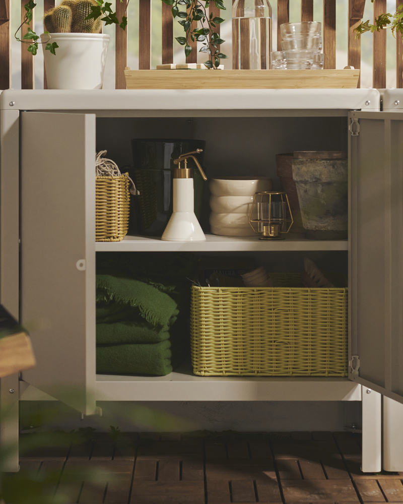 A balcony with a beige KOLBJÖRN cabinet displaying blankets, gardening tools and storage baskets in its open shelves.