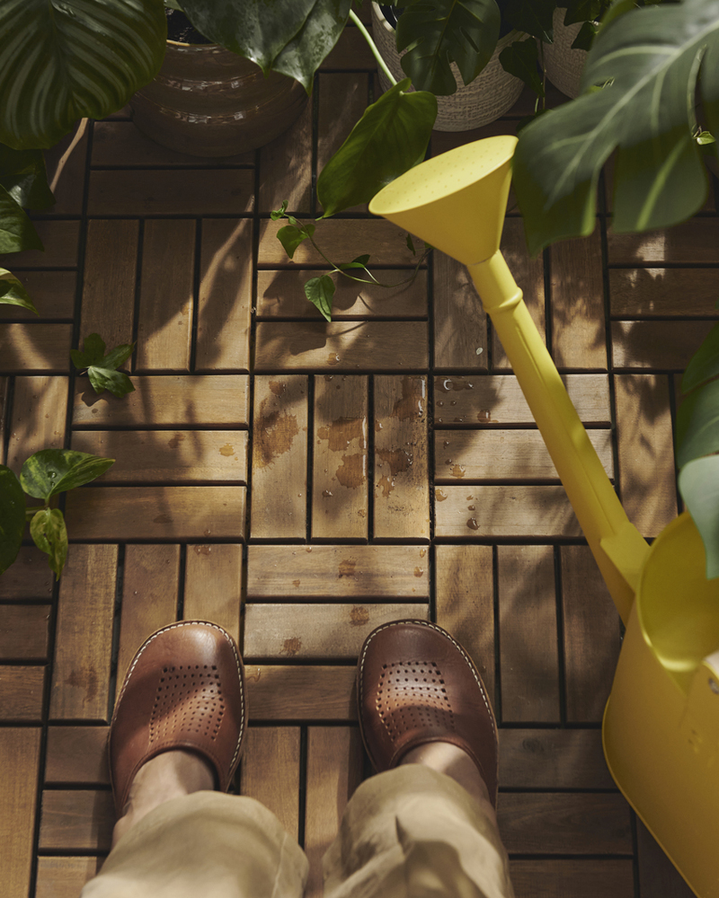 A balcony with acacia RUNNEN outdoor floor decking and a pair of shoes, surrounded by green plants and a yellow watering can.