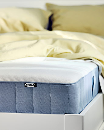 Detalle de la esquina de un colchón azul claro con etiqueta de IKEA, sobre una estructura de cama blanca.