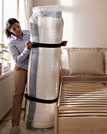 Una mujer sostiene un colchón enrollado y embalado al vacío junto a una estructura de cama de madera con somier.