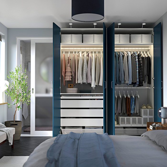 Open PAX wardrobe with blue doors, illuminated interior with hanging clothes, white drawers, and storage boxes in a cozy, organized bedroom.