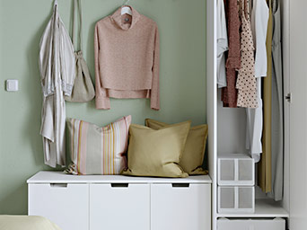 Banco con almacenaje blanco y cojines, junto a ropa colgada en una pared verde suave y un armario abierto con prendas ordenadas.