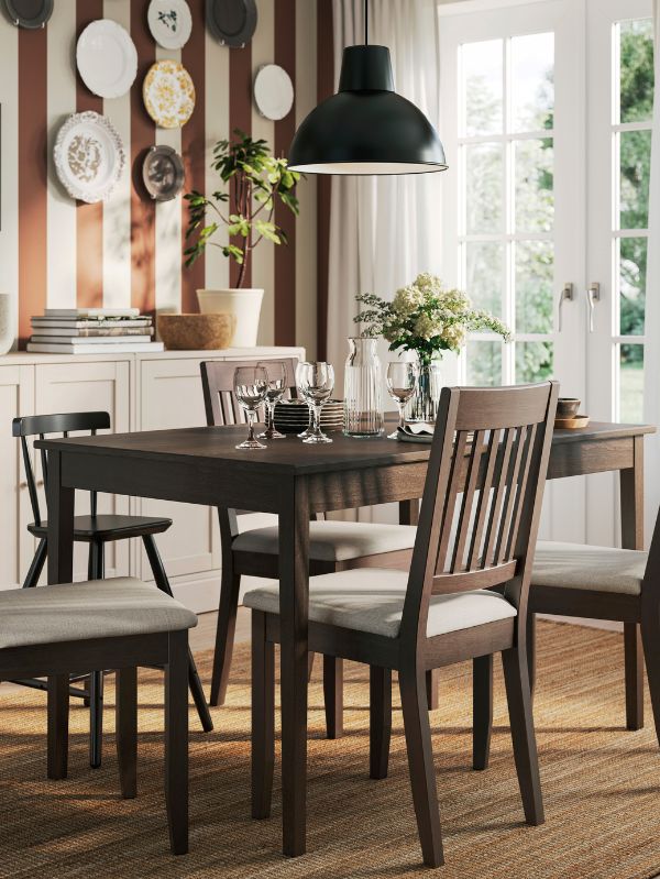 Comedor cálido con lámpara SKURUP, mesa NÄSINGE, alfombra LOHALS, copas SVALKA, jarra VARDAGEN, vajilla GLADELIG y maceta SOJABÖNA.