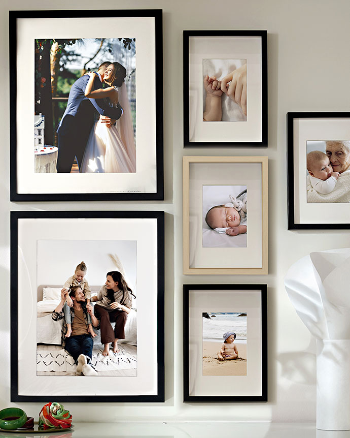 Composición de marcos negros y de madera clara en una pared blanca, mostrando fotos familiares, un bebé durmiendo y una pareja de recién casados.