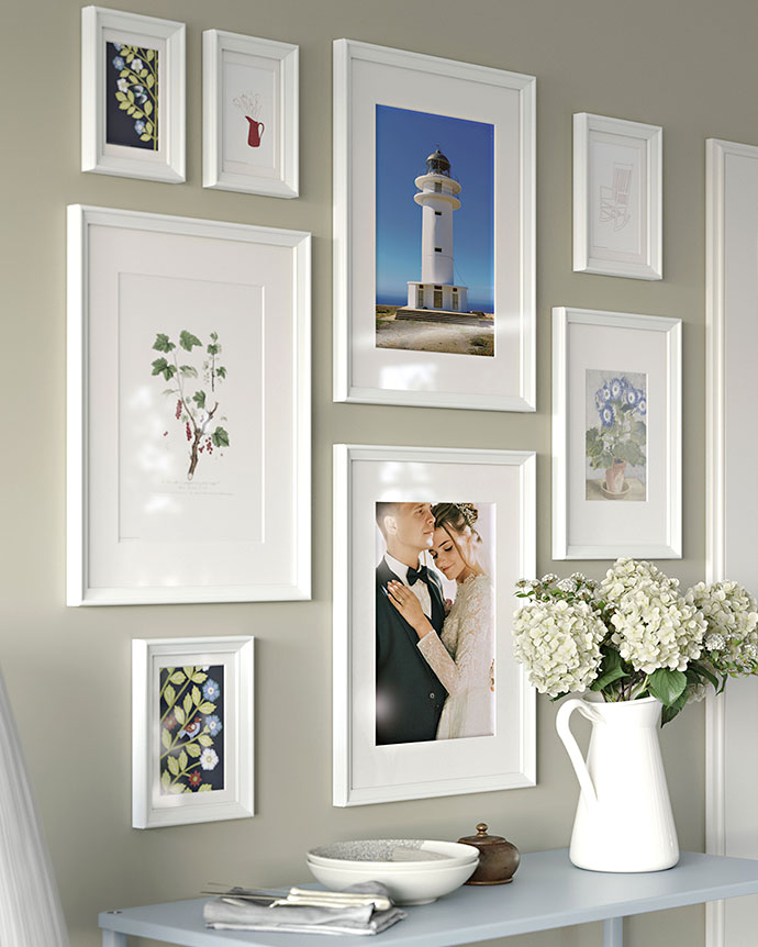 Galería de marcos blancos de distintos tamaños con láminas botánicas, un faro y una foto de boda sobre un aparador azul con flores y cerámica.