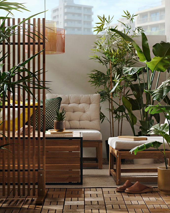 Wooden outdoor furniture with white cushions on a balcony filled with green plants, creating a cozy and natural sunny corner.
