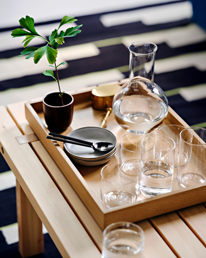 Bandeja de madera sobre mesa con jarra de vidrio, vasos, platos pequeños y una planta joven en maceta oscura, sobre una alfombra de rayas.
