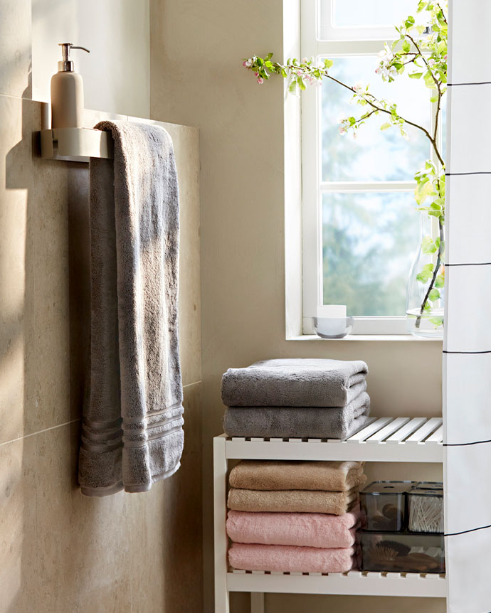 Baño luminoso con toallas dobladas en estante blanco, una toalla colgada, dispensador de jabón y una rama con flores frescas junto a la ventana.