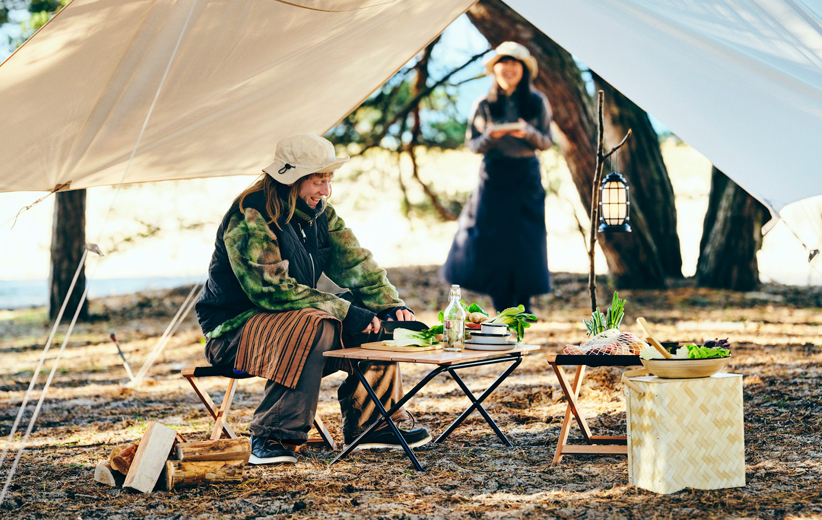 Dos personas sonrientes están preparando comida con artículos de la colección SOLUPPGÅNG en un bosque; están acampando.