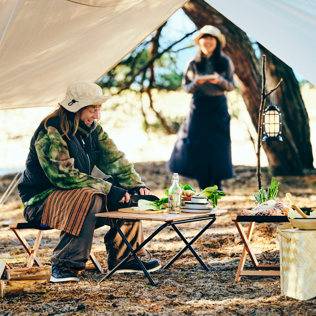 Dos personas sonrientes están preparando comida con artículos de la colección SOLUPPGÅNG en un bosque; están acampando.