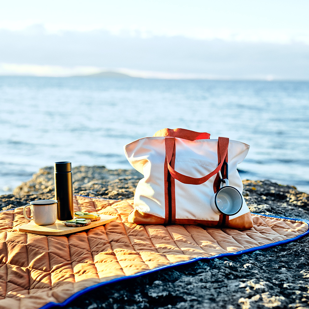 Una bolsa SOLUPPGÅNG, dos tazas y una tabla de cortar están sobre una manta de picnic SOLUPPGÅNG en un acantilado rocoso junto al mar.