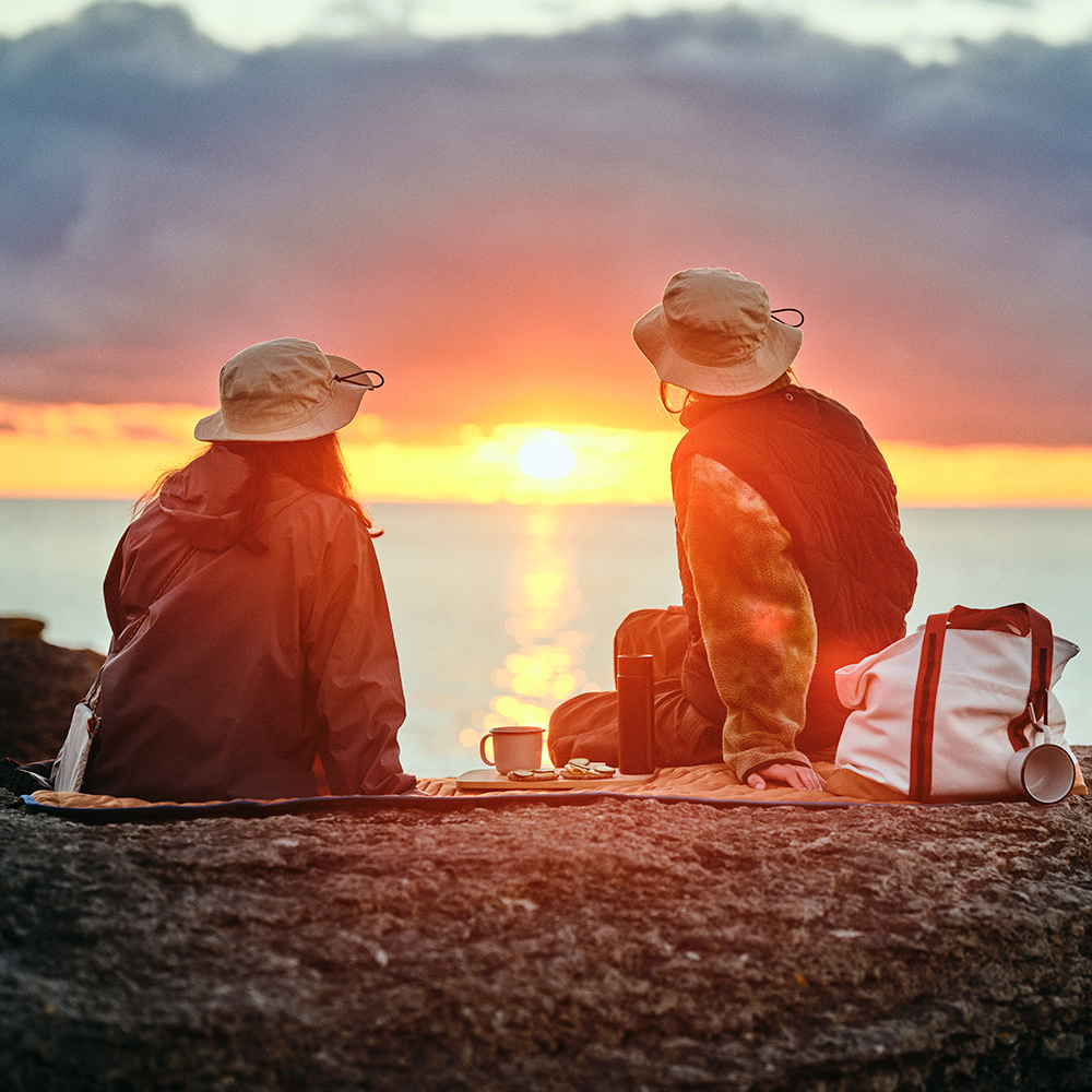 Dos personas que llevan sombreros SOLUPPGÅNG están sentadas sobre una manta de pícnic SOLUPPGÅNG mientras contemplan la puesta de sol junto al mar.