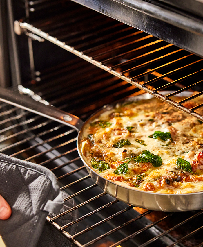 Sartén con comida gratinada y espinacas saliendo del horno, sostenida por una mano con guante de cocina.