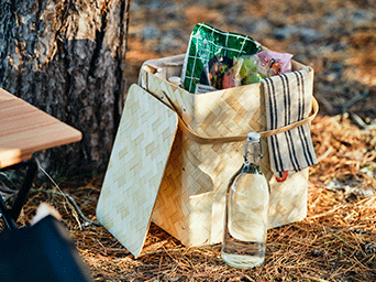 Woven light wood picnic basket filled with snacks and a striped cloth, placed on forest ground next to a glass water bottle.