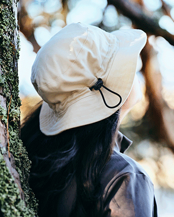 Close-up of a person wearing a beige water-resistant sun hat with an adjustable toggle, leaning against a mossy tree trunk in soft sunlight.