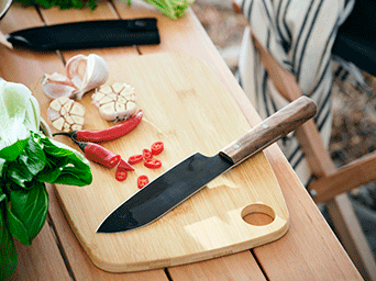 Black chef's knife with a wooden handle on a bamboo cutting board with sliced red chilies, garlic, and fresh green herbs.