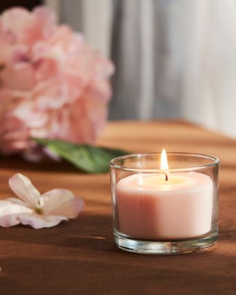 A close-up of a pale pink LOTSFÅGEL scented candle in glass placed on a table with pink flowers in the background