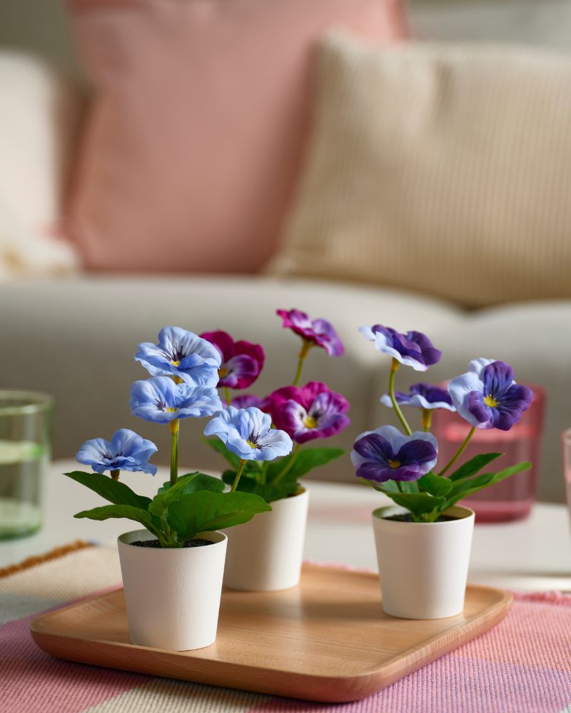 Living room, soft and colorful with PAPPERSBJÖRK glasses, STRANDFLOKA placemat, ÅSVEIG and LAGERPOPPEL cushions, and FEJKA plants.