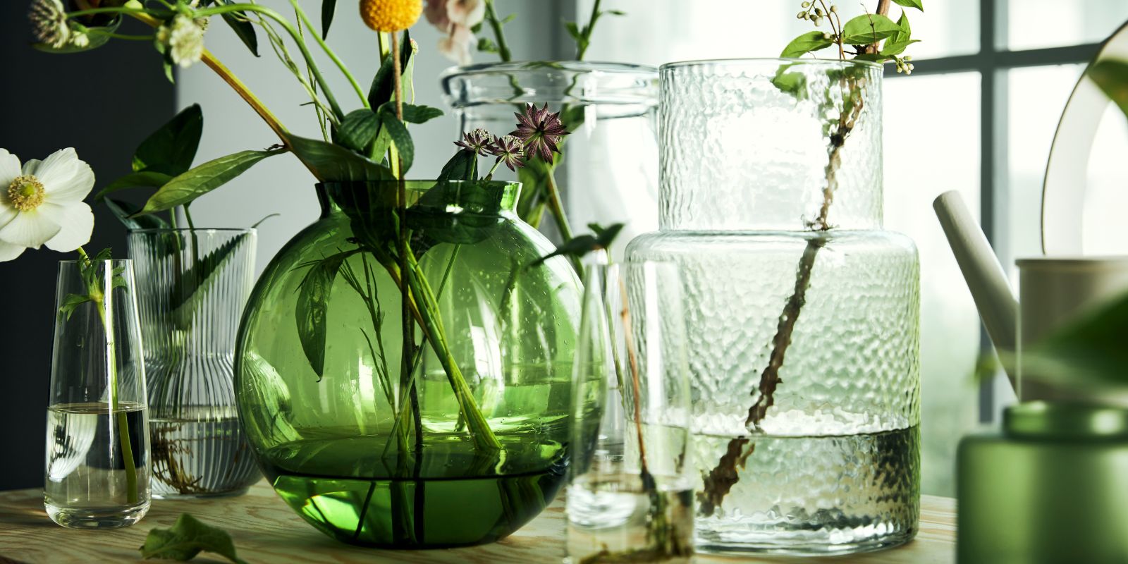 Mesa con jarrones, con PÅDRAG, KONSTFULL, BEGÄRLIG y floreros de vidrio BERÄKNA creando una composición luminosa.