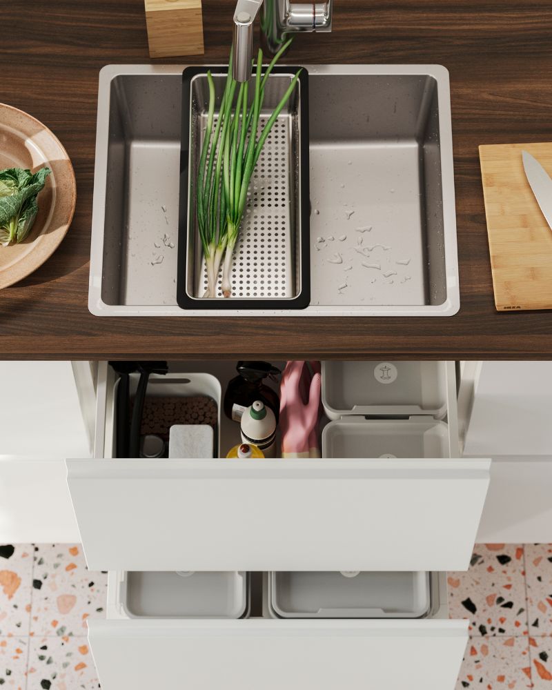 A VRESJÖN inset sink in stainless steel has a NORRSJÖN colander with some spring onions and next to it is a chopping board.