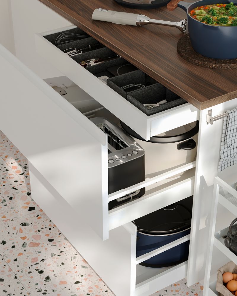Several drawers in a kitchen peninsula with white VOXTORP fronts have been pulled out, revealing small appliances inside.