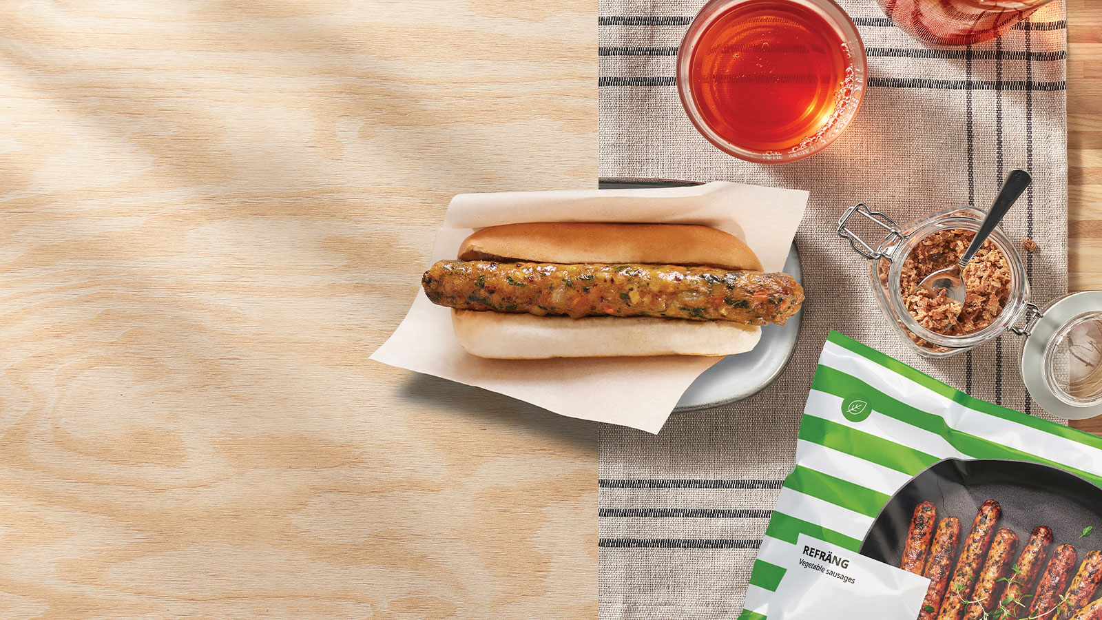 A vegetable sausage in a bun sits on a plate next to a packet of REFRÄNG sausages, a glass of juice, and a jar of crispy onions on a wooden table.