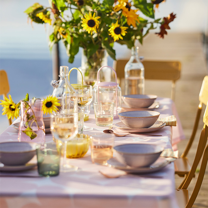 Mesa veraniega al aire libre decorada con flores amarillas, vajilla pastel y cristalería bajo una luz cálida y acogedora.