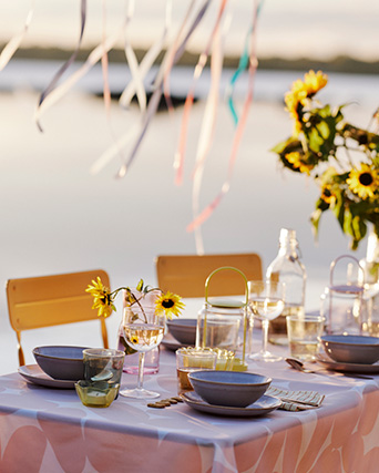 Mesa festiva junto al agua con girasoles, vajilla y cintas de colores bajo una luz suave.