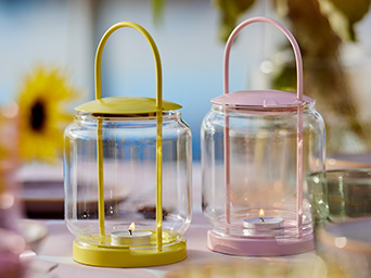 Dos farolillos de cristal, uno amarillo y otro rosa, con velas encendidas sobre una mesa acogedora.