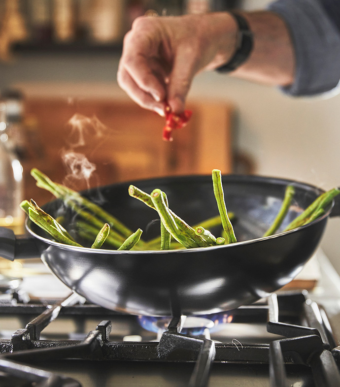 Mano añadiendo chiles a judías verdes en un wok caliente sobre el fuego.