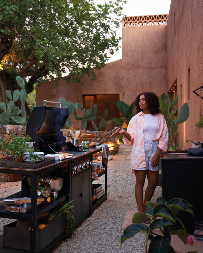 A woman stands in an outdoor courtyard cooking at a grill, surrounded by plants and a modern adobe-style house, holding tongs while preparing food.