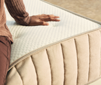 Hand pressing a beige mattress with a textured surface and quilted side panels.