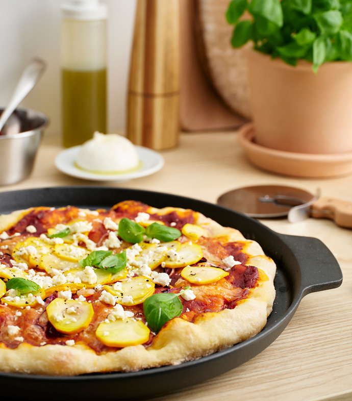 Zucchini and cheese pizza on a black pan, with basil and kitchen utensils in the background.