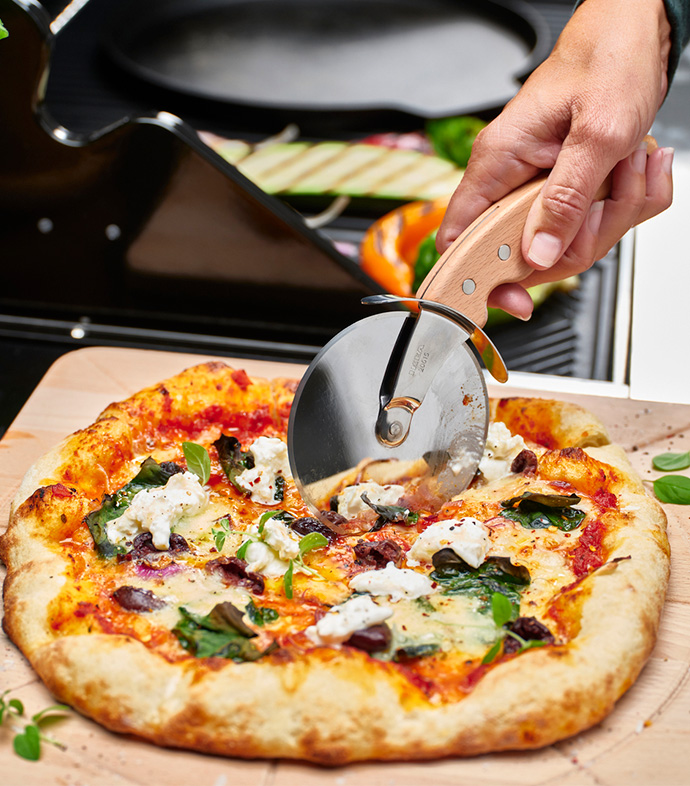 Hand cutting an artisan pizza with a wooden pizza cutter on a wooden board.