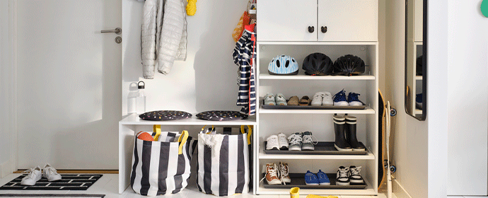 Bright hallway with white modular storage, including a tall cabinet, shoe racks with various footwear and helmets, a bench with striped laundry bags, and a wall-mounted coat rack.