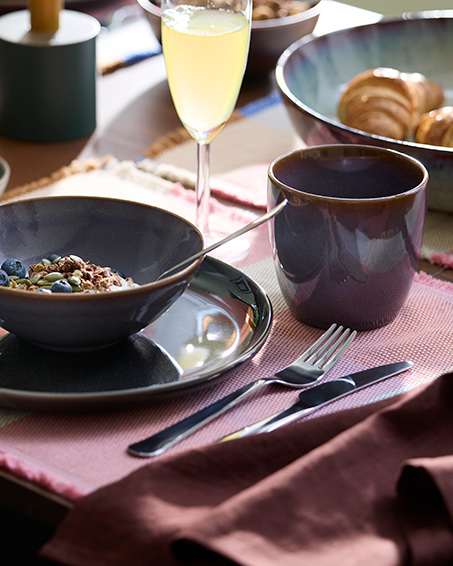 Un cuenco con yogur, muesli y arándanos está colocado junto a una taza GLADELIG de color lila y una copa de champán.