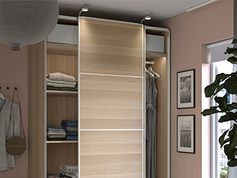 Banco con almacenaje blanco y cojines, junto a ropa colgada en una pared verde suave y un armario abierto con prendas ordenadas.