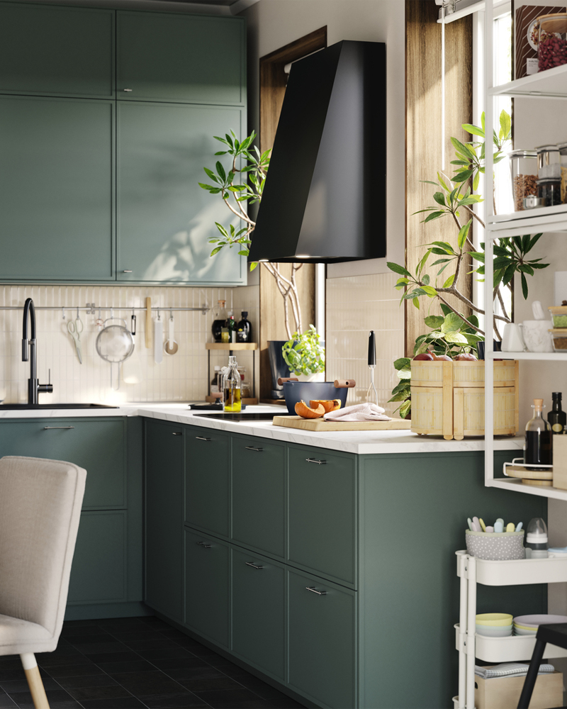 Una cocina pequeña cuenta con varios armarios bajos y altos con frentes ASPUDDEN de color verde grisáceo y una campana extractora junto a una ventana.