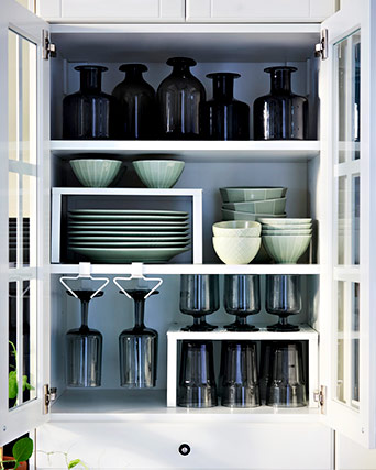 Armario blanco abierto con vajilla verde suave, copas colgadas y botes de vidrio oscuro, todo perfectamente organizado con estantes adicionales.